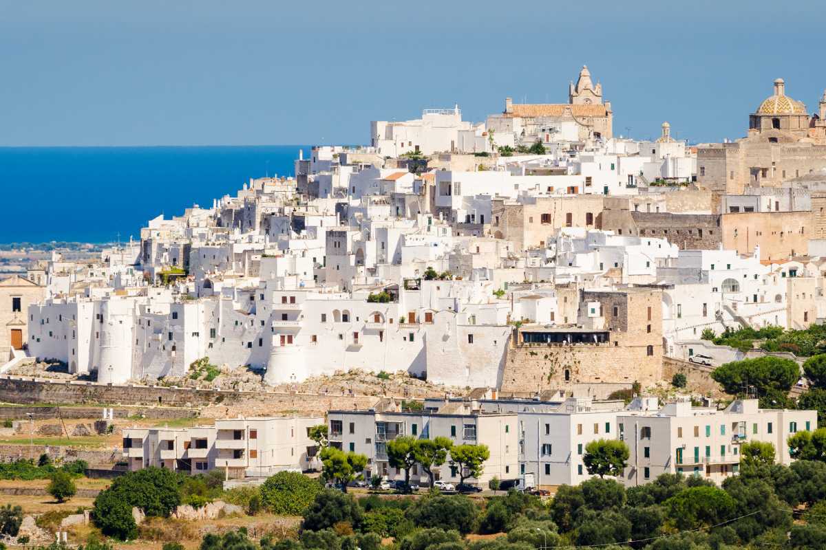 Vista su auto parcheggiate vicino alle mura di Ostuni, con turisti che camminano verso il centro bianco, atmosfera estiva